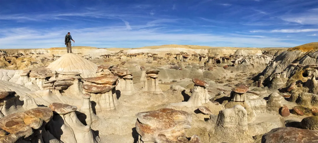 A backpacker with trekking poles stands on a pale sandstone dome, looking out across a desert of hoodoos under a blue sky.